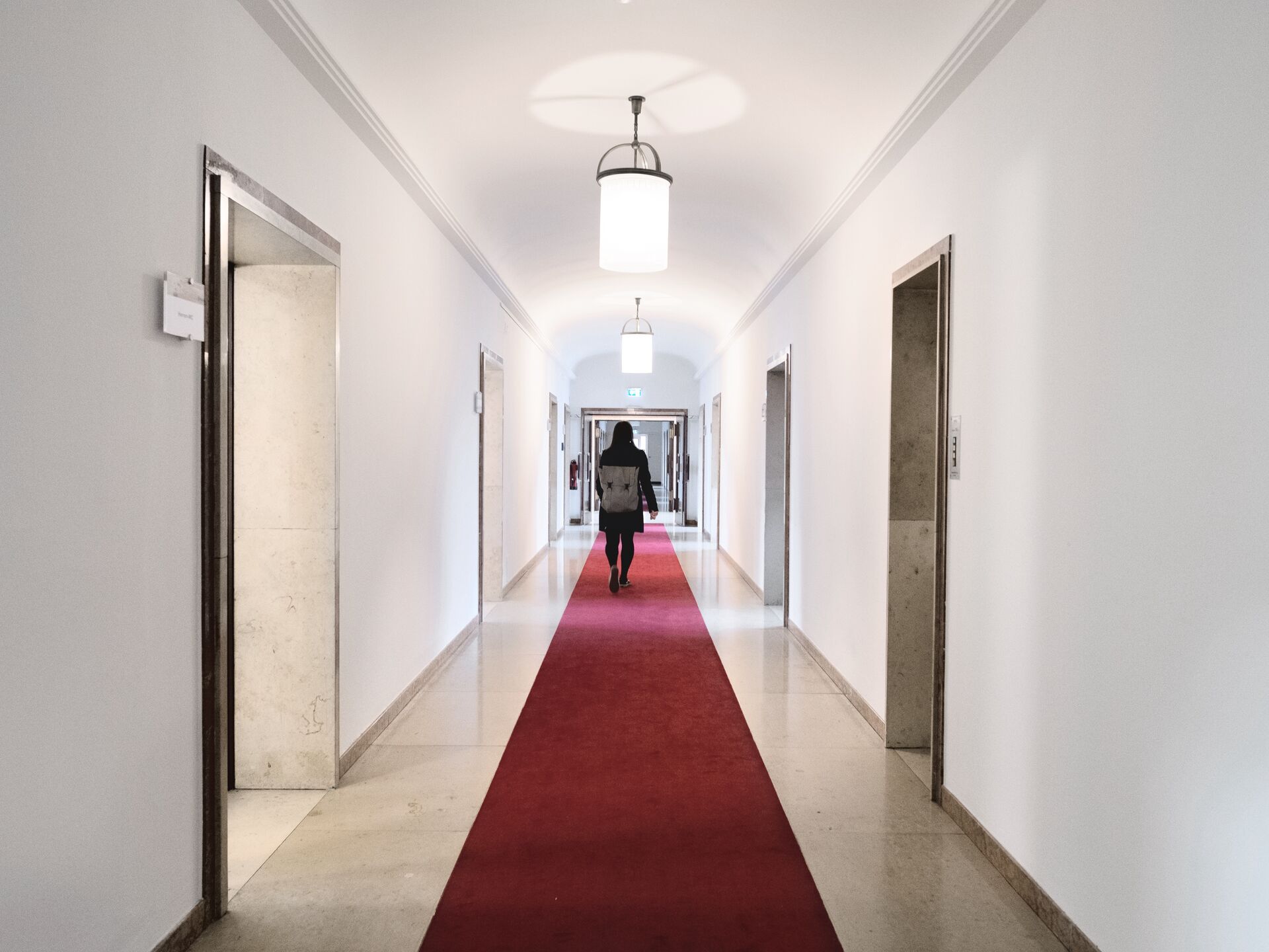 Person geht durch einen hellen Flur mit rotem Teppich und modernen Lampen, Blickrichtung zum Ausgang.