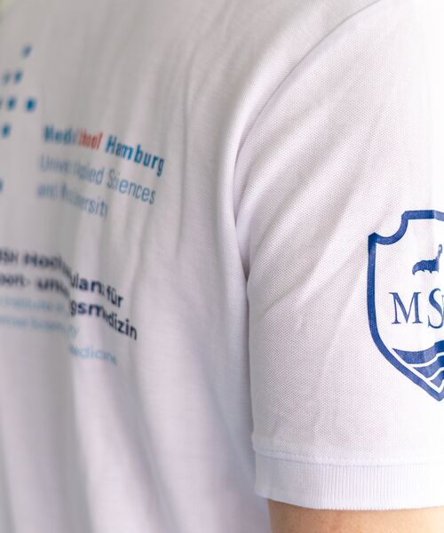 Rückansicht einer Person im weißen T-Shirt mit MSH-Logo und Schriftzug „Medical School Hamburg“.
