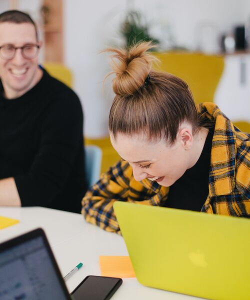 Zwei Personen sitzen lachend an einem Tisch mit Laptops in einer modernen Arbeitsumgebung.