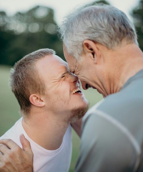 Ein junger Mann und ein älterer Mann berühren lachend die Stirn aneinander und zeigen Zuneigung.
