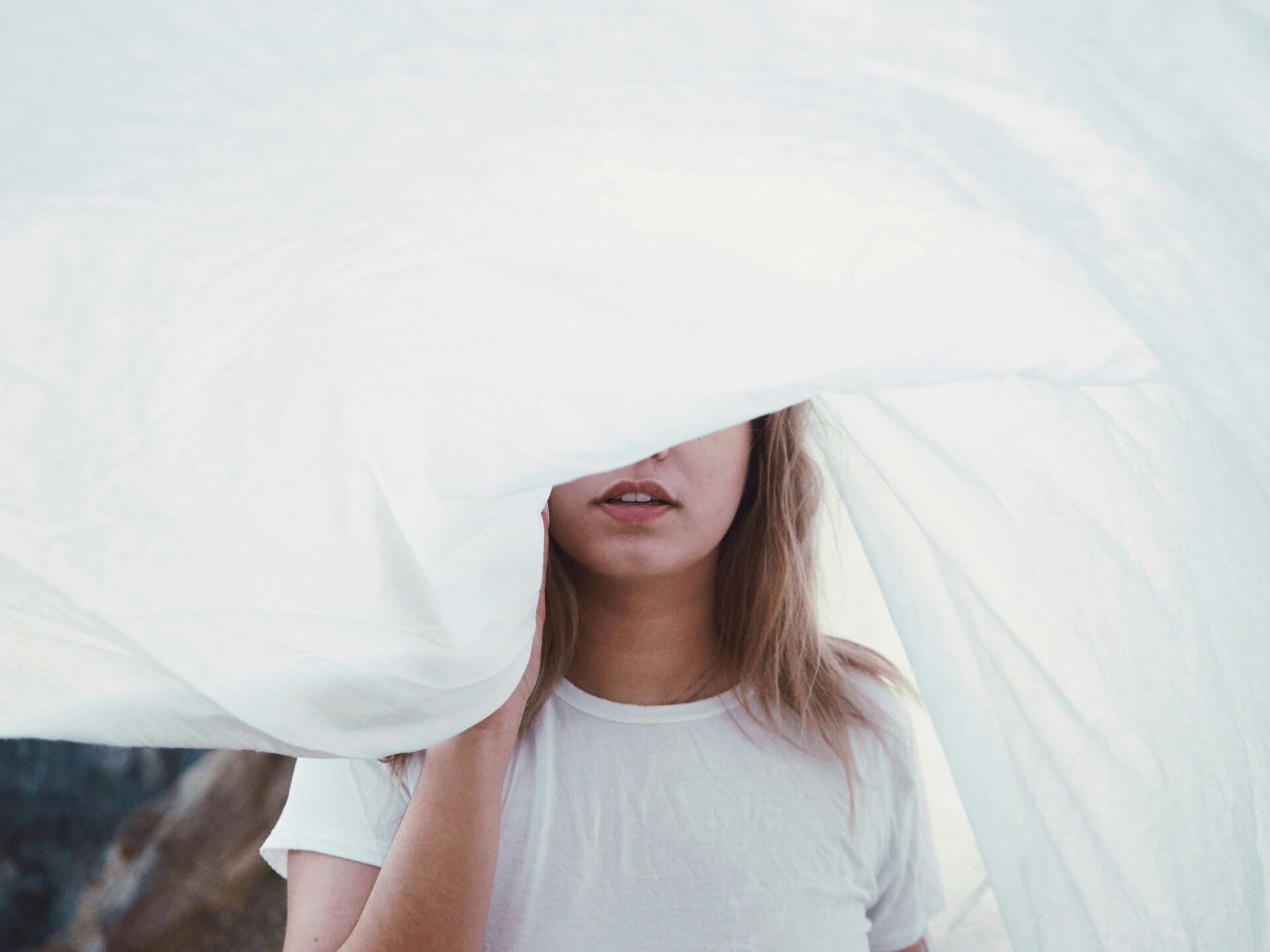 Person in weißem T-Shirt steht hinter einem wehenden, halbtransparenten Tuch, das ihr Gesicht teilweise verdeckt.