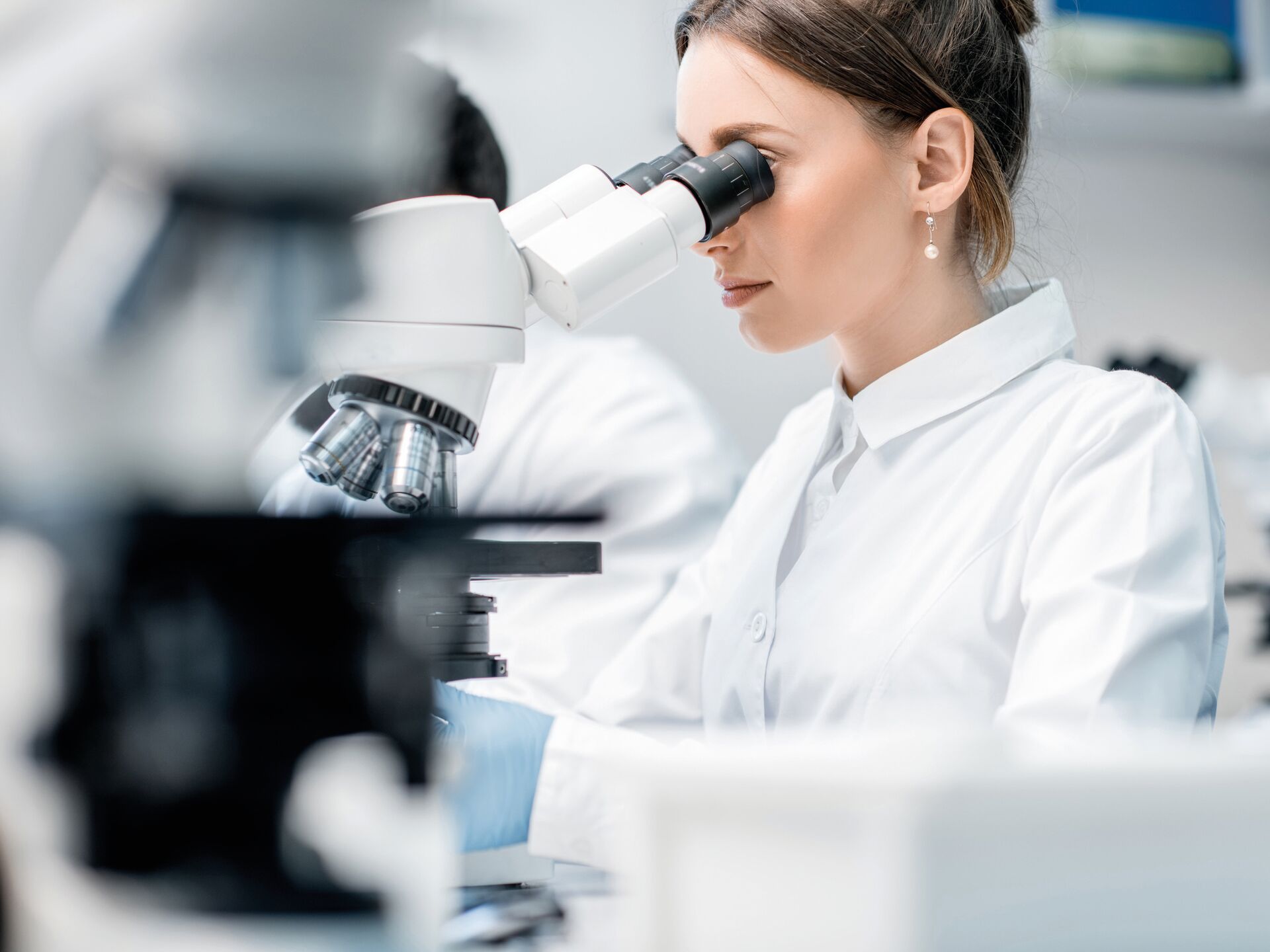 Eine Forscherin im weißen Laborkittel betrachtet eine Probe durch ein Mikroskop.