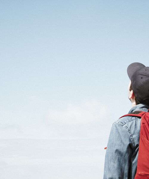 Person mit rotem Rucksack und Kappe blickt in den blauen Himmel, Symbol für Freiheit und neue Perspektiven.