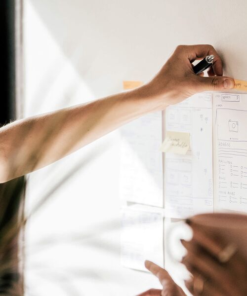 Person schreibt Notizen auf ein Whiteboard mit Skizzen und Post-its, vermutlich bei einer Teamarbeit oder Projektplanung.