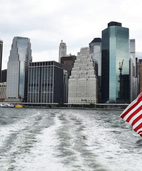 Blick vom Wasser auf eine Skyline mit hohen Bürogebäuden, am rechten Bildrand eine wehende US-Flagge.