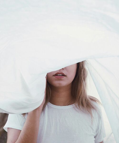 Person in weißem T-Shirt steht hinter einem wehenden, halbtransparenten Tuch, das ihr Gesicht teilweise verdeckt.