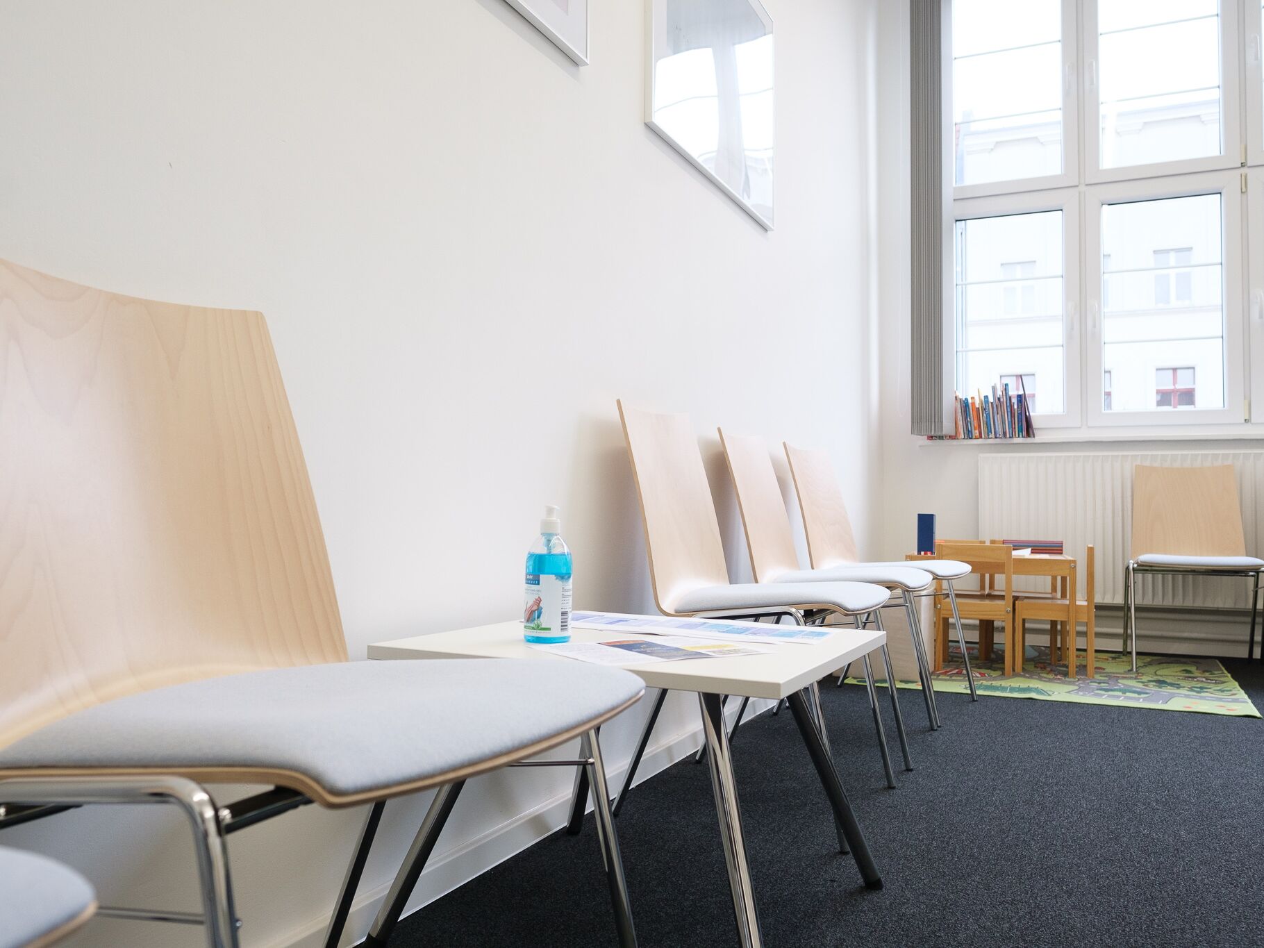 Heller Warteraum mit modernen Holzstühlen, einem kleinen Tisch mit Desinfektionsmittel und Zeitschriften.