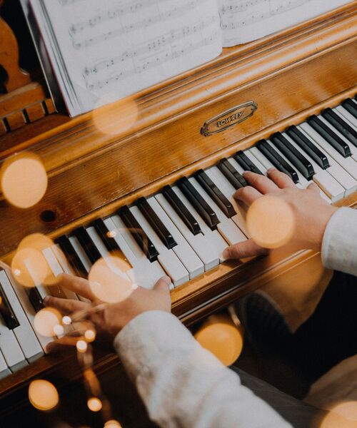 Person spielt auf einem Holzklavier, mit Notenblatt im Hintergrund und warmen Lichtreflexen im Vordergrund.
