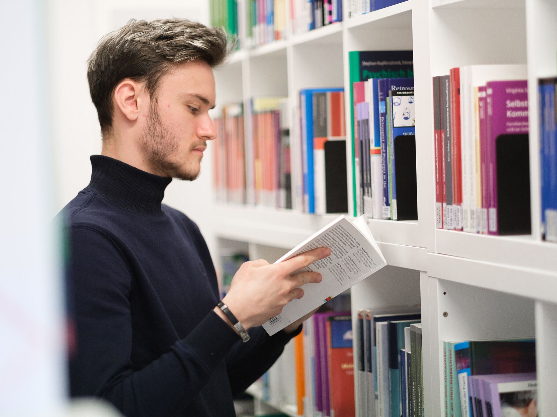 Person steht vor einem Bücherregal und liest in einem Buch, umgeben von Fachliteratur.