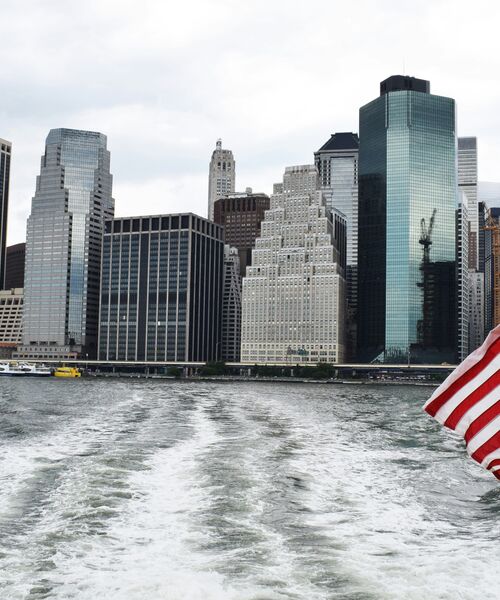 Blick vom Wasser auf eine Skyline mit hohen Bürogebäuden, am rechten Bildrand eine wehende US-Flagge.