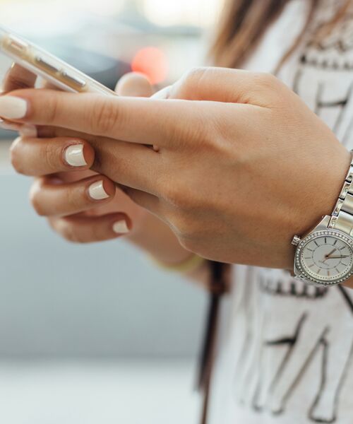 Nahaufnahme einer Person, die ein Smartphone in den Händen hält, mit silberner Armbanduhr und dezent lackierten Fingernägeln.