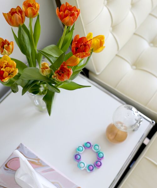 Ein heller Tisch mit orangefarbenen Tulpen, einer Lampe und kleinen bunten Ringen neben einem cremefarbenen Ledersessel.