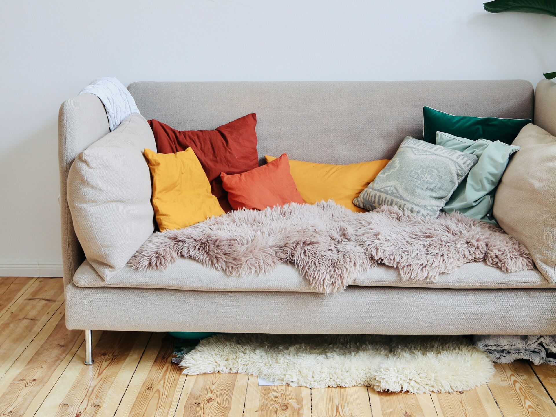 Beiges Sofa mit bunten Kissen und Decke in gemütlich eingerichteter Wohnumgebung mit Holzboden und Pflanze.