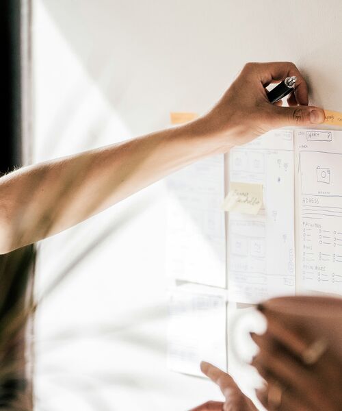 Person schreibt Notizen auf ein Whiteboard mit Skizzen und Post-its, vermutlich bei einer Teamarbeit oder Projektplanung.