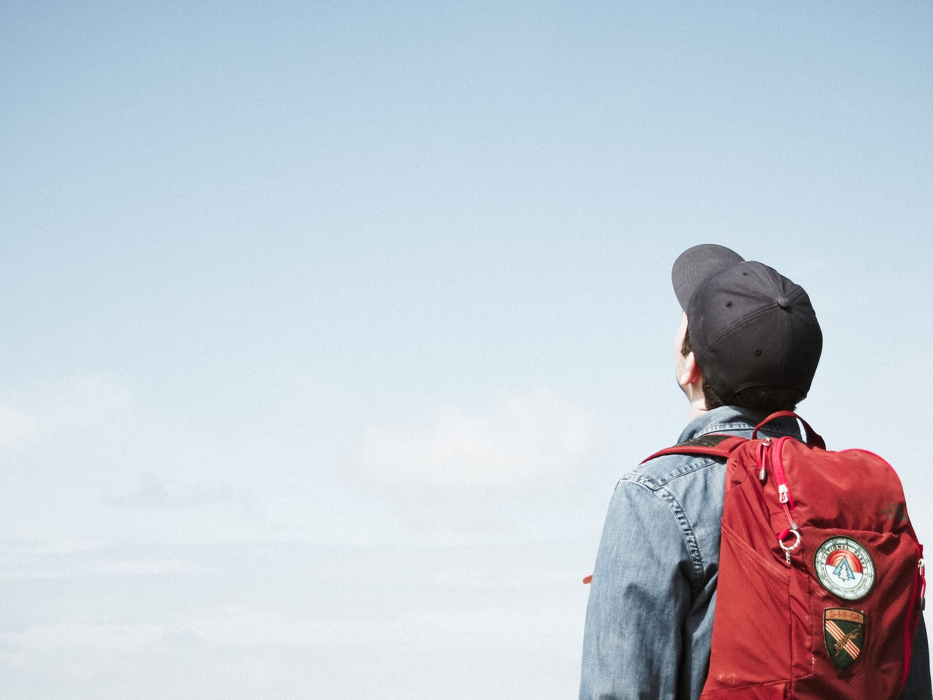 Person mit rotem Rucksack und Kappe blickt in den blauen Himmel, Symbol für Freiheit und neue Perspektiven.