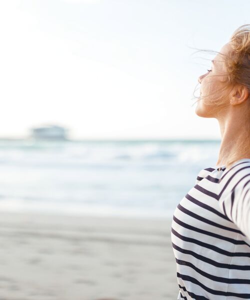 Eine Frau steht am Strand, schließt die Augen und atmet tief ein, während der Wind durchs Haar weht.