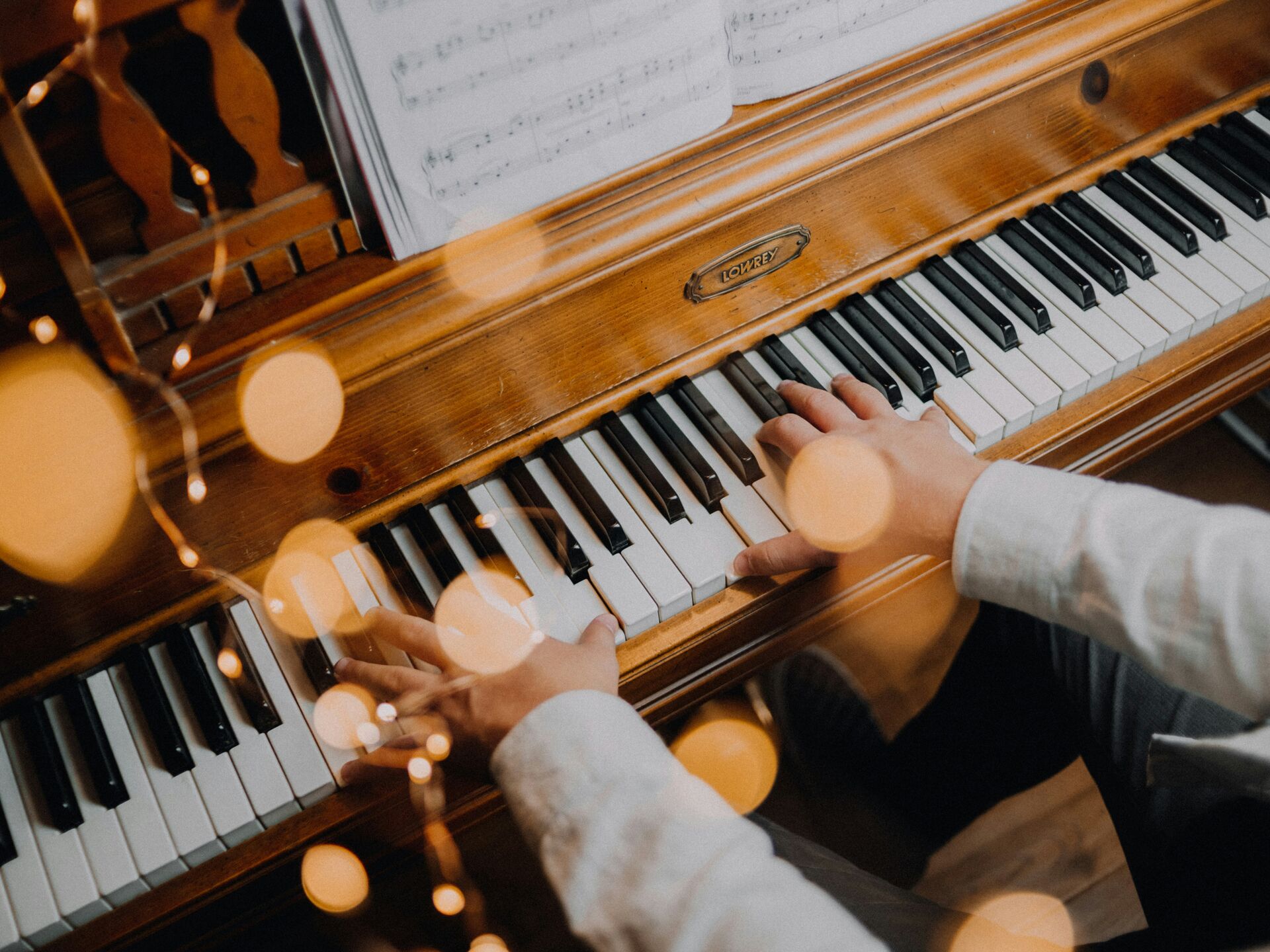 Person spielt auf einem Holzklavier, mit Notenblatt im Hintergrund und warmen Lichtreflexen im Vordergrund.