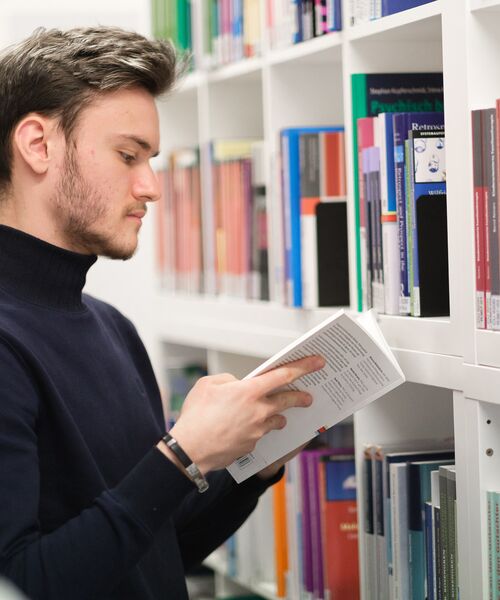 Person steht vor einem Bücherregal und liest in einem Buch, umgeben von Fachliteratur.