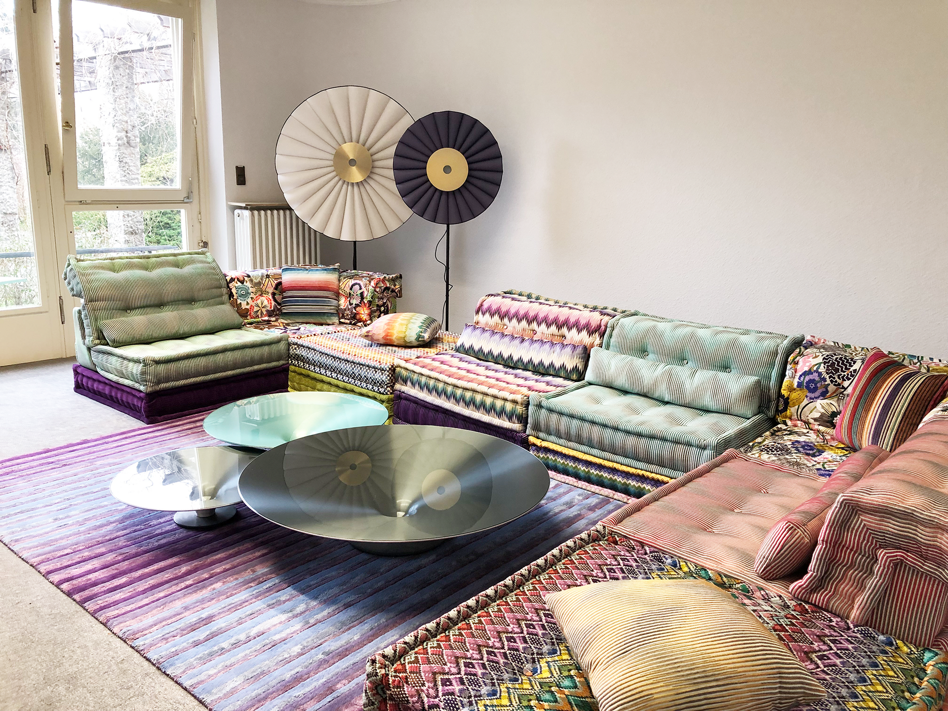 Wohnzimmer mit bunten, gemusterten Sofas, runden Couchtischen und zwei großen, dekorativen Stehlampen im Hintergrund.