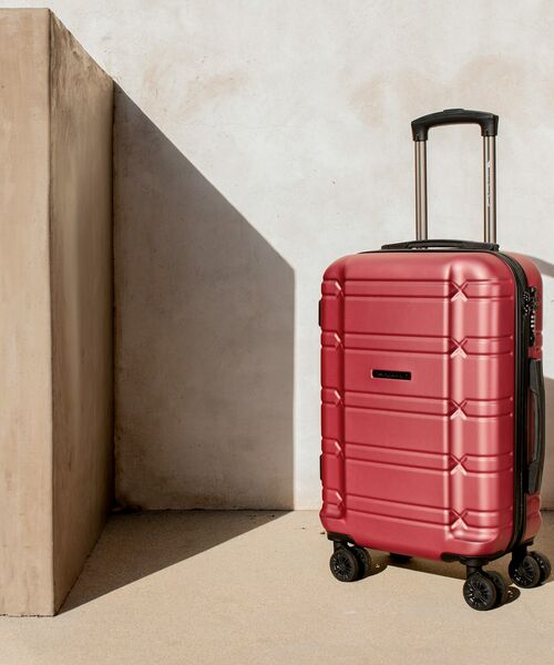 Roter Hartschalenkoffer mit ausziehbarem Griff steht in hellem Sonnenlicht vor beiger Wand.