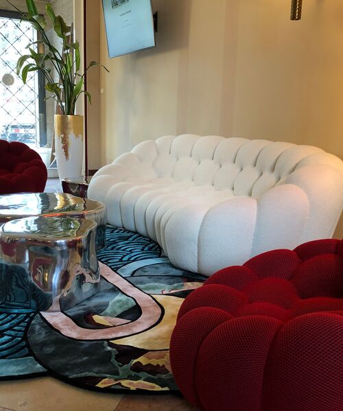 Modern eingerichtete Sitzecke mit weißem Sofa, roten Sesseln und einem silbernen Couchtisch auf buntem Teppich.