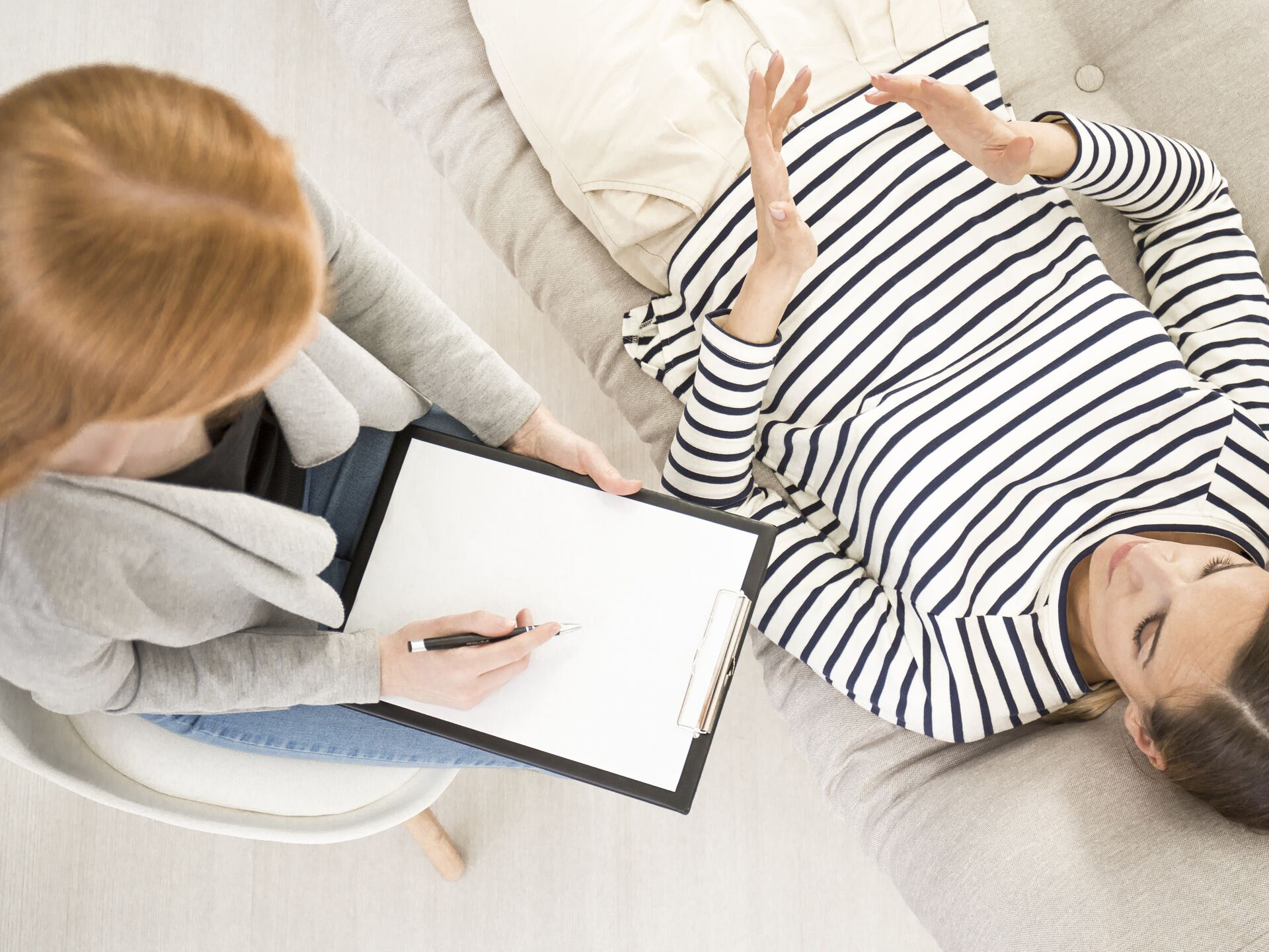 Beratungssituation, bei der eine Person auf einem Sofa liegt und mit einer Fachperson spricht, die Notizen auf einem Klemmbrett macht.