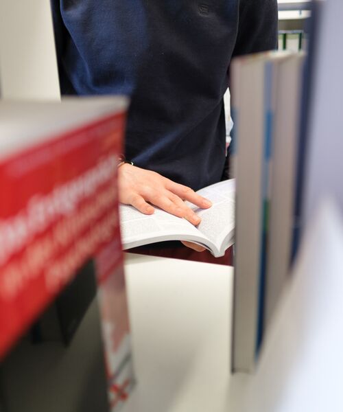 Person blättert in einem aufgeschlagenen Buch zwischen Bücherregalen in einer Bibliothek.