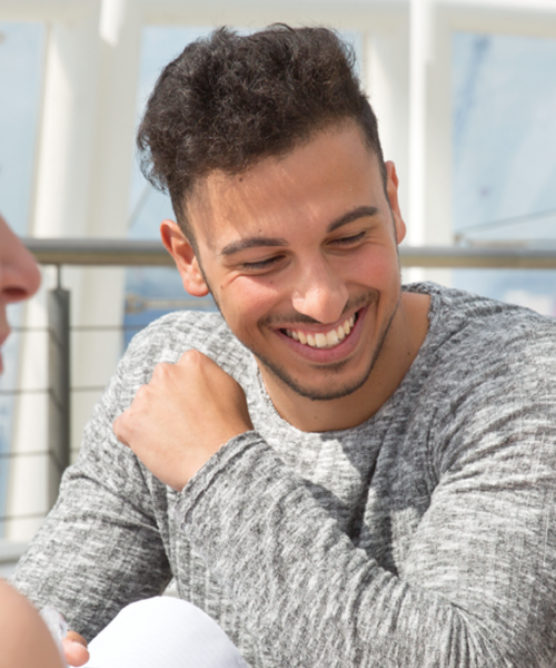 Junger Mann sitzt in heller Umgebung und lacht herzlich im Gespräch mit einer anderen Person.