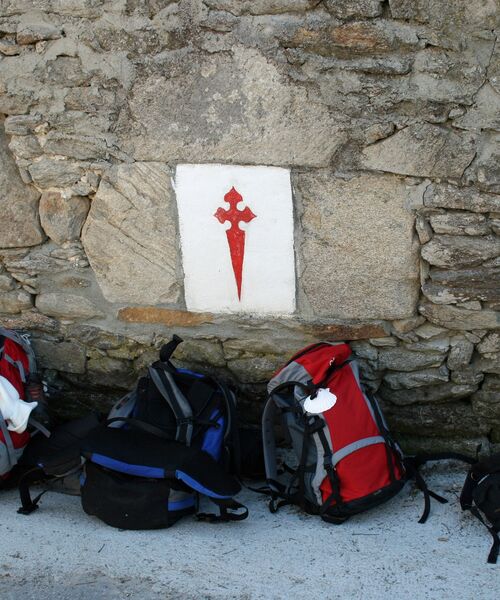 Mehrere Rucksäcke lehnen an einer Steinmauer mit dem roten Jakobskreuz, Symbol des Jakobswegs.