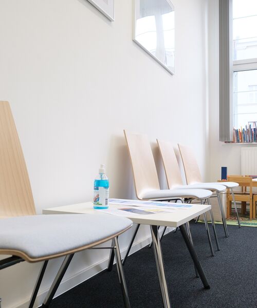 Heller Warteraum mit modernen Holzstühlen, einem kleinen Tisch mit Desinfektionsmittel und Zeitschriften.