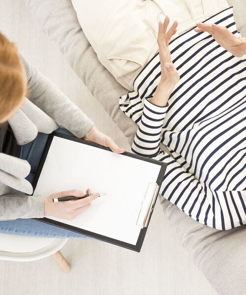 Beratungssituation, bei der eine Person auf einem Sofa liegt und mit einer Fachperson spricht, die Notizen auf einem Klemmbrett macht.