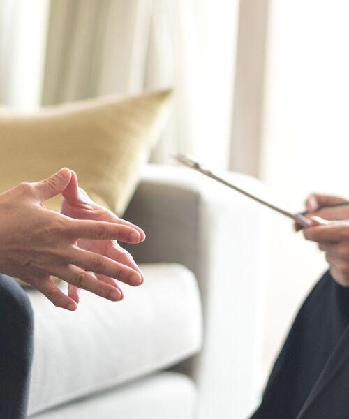 Zwei Personen im Gespräch auf einem Sofa, eine Person spricht mit gestikulierenden Händen, die andere hält einen Schreibblock.