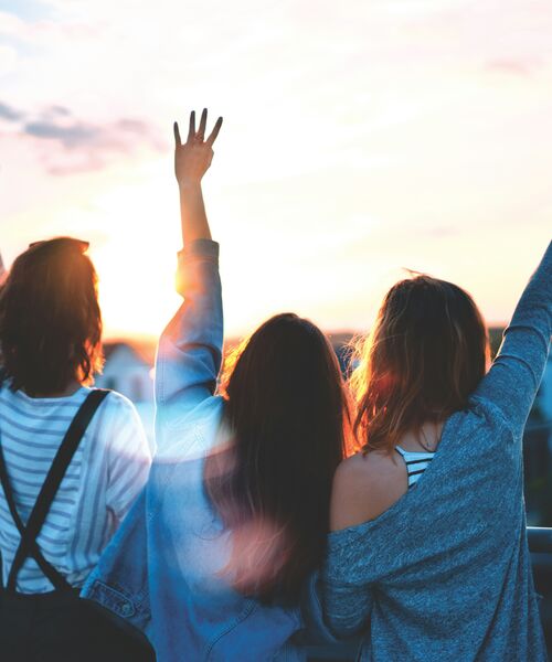 Drei Personen stehen nebeneinander im Sonnenuntergang, zwei heben die Arme fröhlich in die Luft.