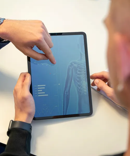 Personen betrachten auf einem Tablet eine digitale Darstellung eines menschlichen Skeletts mit Diagrammen und Daten.