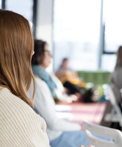 Mehrere Personen sitzen in einem Raum und hören aufmerksam zu, im Vordergrund ist unscharf der Hinterkopf einer Frau mit langen, hellbraunen Haaren zu sehen.
