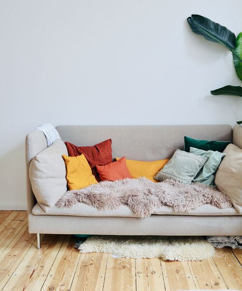 Beiges Sofa mit bunten Kissen und Decke in gemütlich eingerichteter Wohnumgebung mit Holzboden und Pflanze.