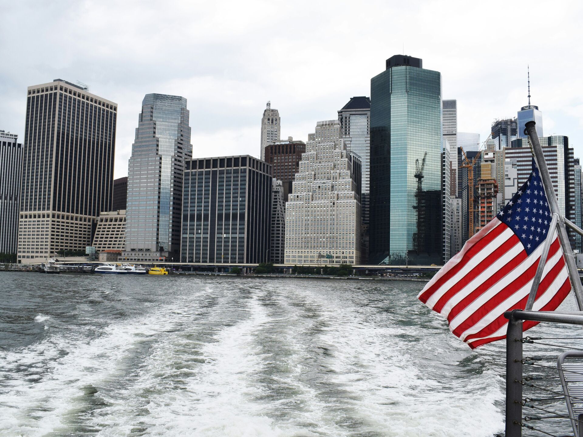 Blick vom Wasser auf eine Skyline mit hohen Bürogebäuden, am rechten Bildrand eine wehende US-Flagge.