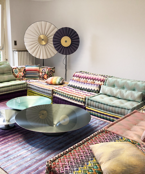 Wohnzimmer mit bunten, gemusterten Sofas, runden Couchtischen und zwei großen, dekorativen Stehlampen im Hintergrund.