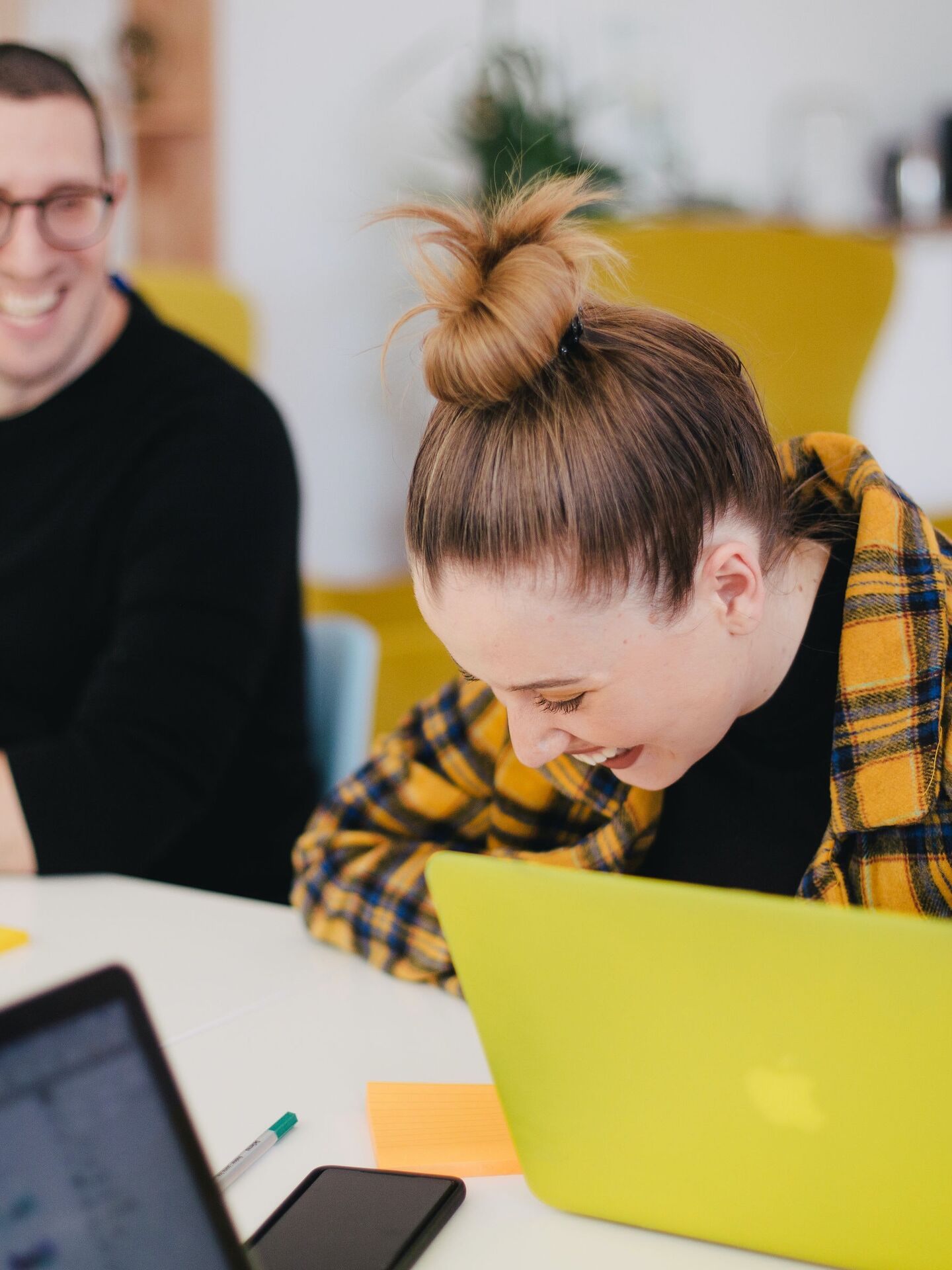 Zwei Personen sitzen lachend an einem Tisch mit Laptops in einer modernen Arbeitsumgebung.