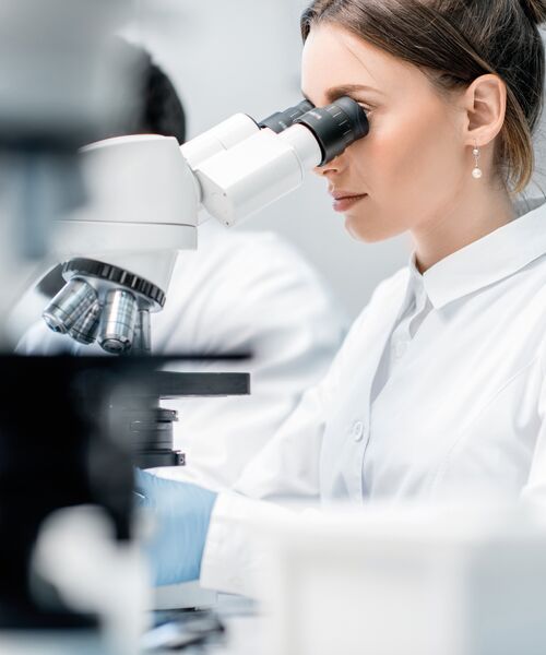 Eine Forscherin im weißen Laborkittel betrachtet eine Probe durch ein Mikroskop.